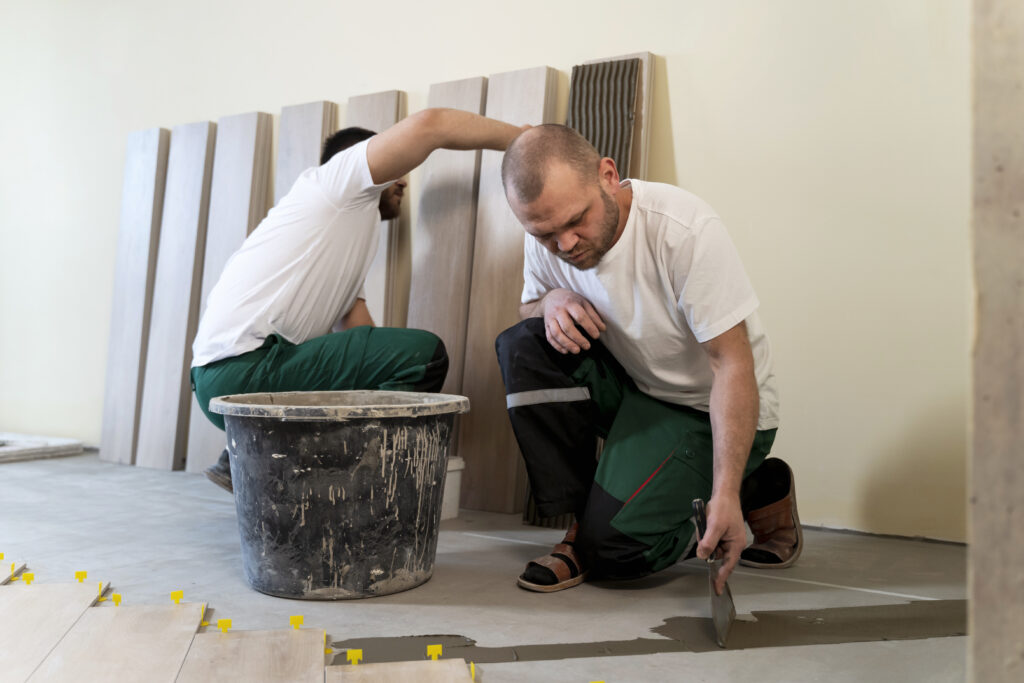 Zwei Handwerker verteilen Kleber auf dem Boden und bereiten Holzdielen zur Verlegung vor.