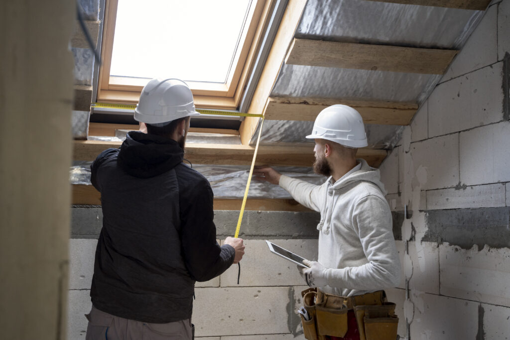 Zwei Handwerker mit weißen Schutzhelmen vermessen gemeinsam ein Dachfenster in einem unausgebauten Dachgeschoss mit sichtbarer Dämmung und Mauerwerk.