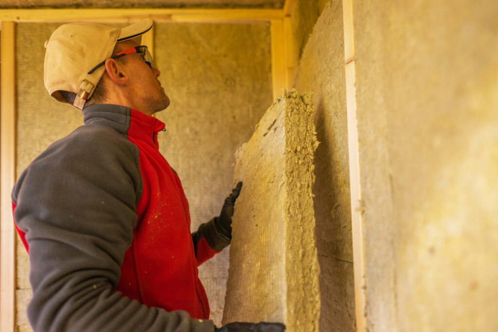 Ein Handwerker in roter Jacke und beigefarbener Kappe installiert Mineralwolldämmung in einem Holzrahmen zur Energieoptimierung.