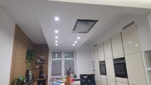 Moderne Küche mit weißen Hochglanzschränken, LED-Deckenspots und einer dekorativen Holzwand mit Lamellen.