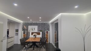 Moderner Essbereich mit Holztisch, grauen Polsterstühlen, LED-Deckenspots und indirekter Beleuchtung entlang der Decke.