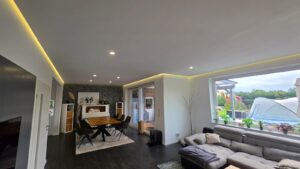 Offener Wohn- und Essbereich mit grauem Ecksofa, Holzesstisch, LED-Einbauspots und warmer indirekter Deckenbeleuchtung.
