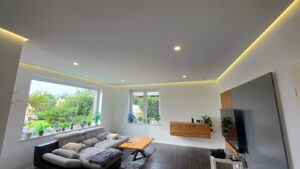 Modernes Wohnzimmer mit grauem Ecksofa, großem Fenster, LED-Deckenspots und warmweißer indirekter Beleuchtung.